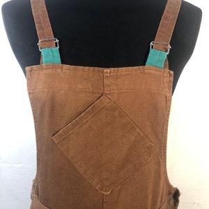 Pode incluir: Macac&atilde;o marrom com al&ccedil;as ajust&aacute;veis e um bolso frontal. As al&ccedil;as s&atilde;o presas com ferragens de metal prateado e apresentam um detalhe em verde-azulado. O macac&atilde;o &eacute; feito de um tecido dur&aacute;vel e tem um design cl&aacute;ssico.
