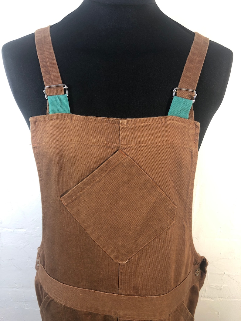 Pode incluir: Macac&atilde;o marrom com al&ccedil;as ajust&aacute;veis e um bolso frontal. As al&ccedil;as s&atilde;o presas com ferragens de metal prateado e apresentam um detalhe em verde-azulado. O macac&atilde;o &eacute; feito de um tecido dur&aacute;vel e tem um design cl&aacute;ssico.