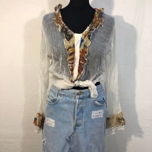 May include: A white sheer blouse with a brown and white patterned ruffle trim around the neckline and cuffs. The blouse is tied at the waist and is worn over a light blue denim skirt with patches.