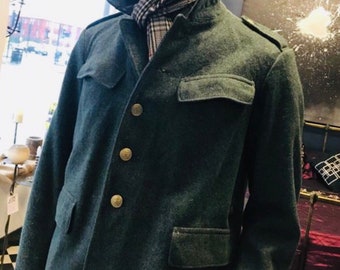 Puede incluir: Una chaqueta de lana verde oscuro con cierre de botones y un bolsillo de solapa en el pecho. La chaqueta tiene cuello y mangas largas. La persona que lleva la chaqueta también lleva una bufanda estampada y gafas de sol.