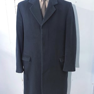 May include: A dark navy blue overcoat with a classic design. The coat features a notched lapel, two front pockets, and a three-button closure. The coat is displayed on a mannequin, revealing a light-colored shirt and patterned tie underneath.