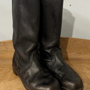 May include: A pair of black leather boots with a tall shaft. The boots have a rounded toe and a flat sole. The leather appears worn, with visible creases and a matte finish. The boots are placed on a wooden surface.