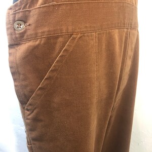 Pode incluir: Close-up de macac&atilde;o marrom. O macac&atilde;o apresenta um bolso frontal grande, um fecho de bot&atilde;o na cintura e uma cintura larga. O tecido parece ser algod&atilde;o resistente ou material semelhante. O design geral &eacute; simples e funcional.