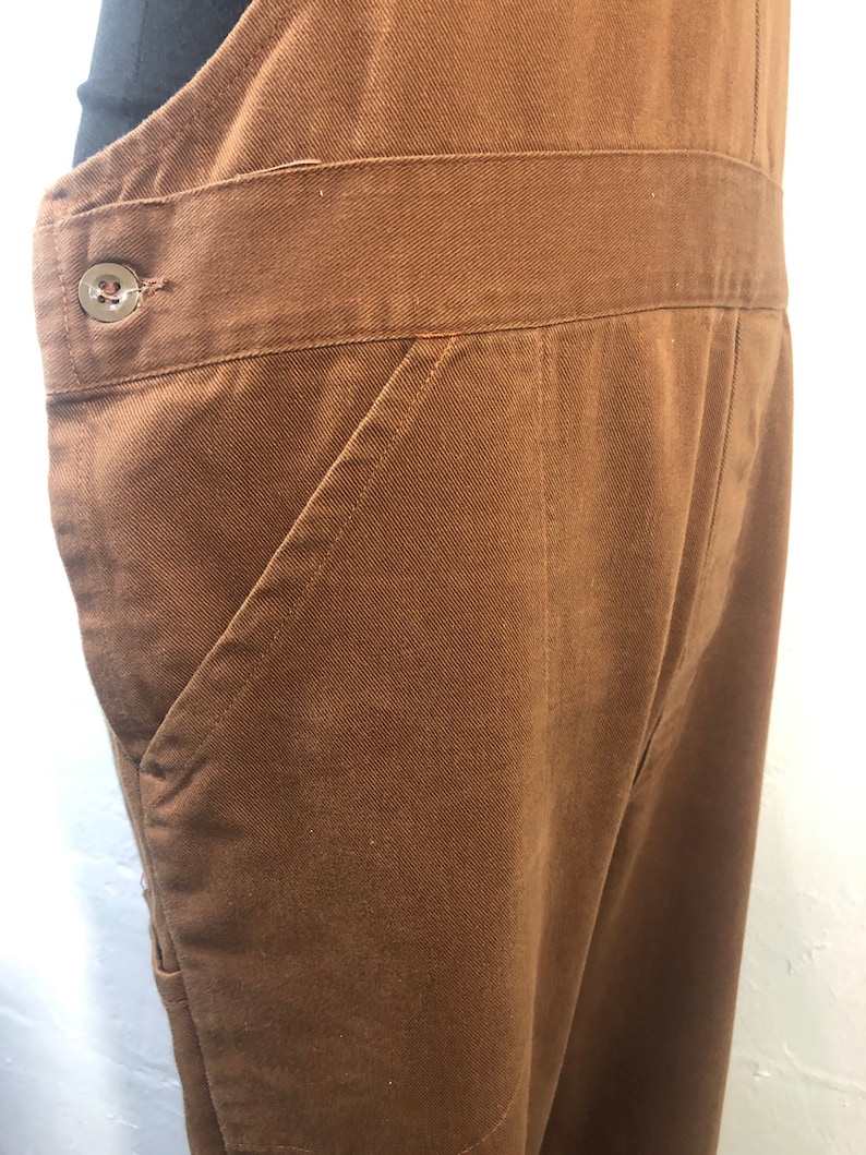 Pode incluir: Close-up de macac&atilde;o marrom. O macac&atilde;o apresenta um bolso frontal grande, um fecho de bot&atilde;o na cintura e uma cintura larga. O tecido parece ser algod&atilde;o resistente ou material semelhante. O design geral &eacute; simples e funcional.