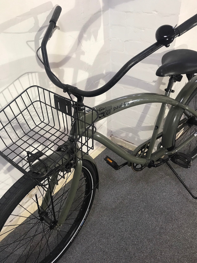 May include: A grey cruiser bicycle with a black wire basket, black handlebars, and a black seat. The bicycle has black tyres with white sidewalls and a bell on the handlebars. The frame has the text "paul frank".