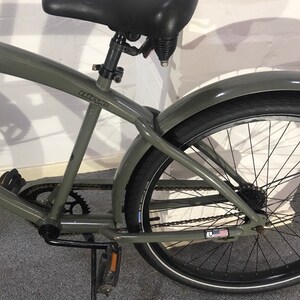 May include: A grey cruiser bicycle with a black saddle, mudguards, and tyres. The bike features a chain, pedals, and rear suspension. The tyres have a white stripe. The bike frame has the word "nine" printed on it.