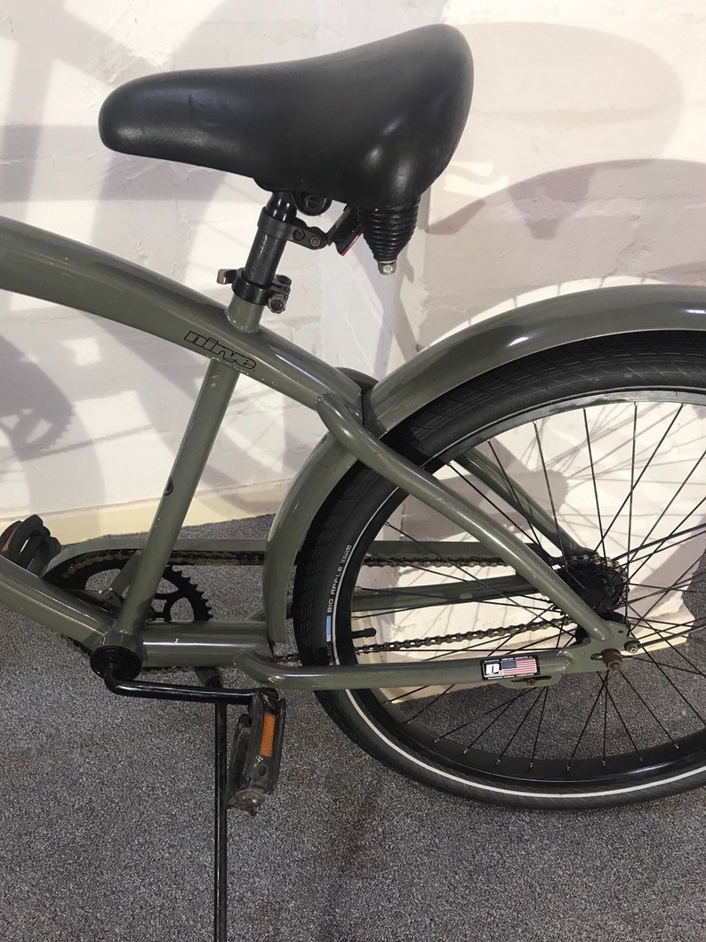 May include: A grey cruiser bicycle with a black saddle, mudguards, and tyres. The bike features a chain, pedals, and rear suspension. The tyres have a white stripe. The bike frame has the word "nine" printed on it.