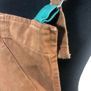 Pode incluir: Close-up de um avental marrom com detalhe de al&ccedil;a verde-azulado e ferragens de metal prateado. O avental tem um bolso e &eacute; feito de um tecido dur&aacute;vel. As al&ccedil;as s&atilde;o ajust&aacute;veis.