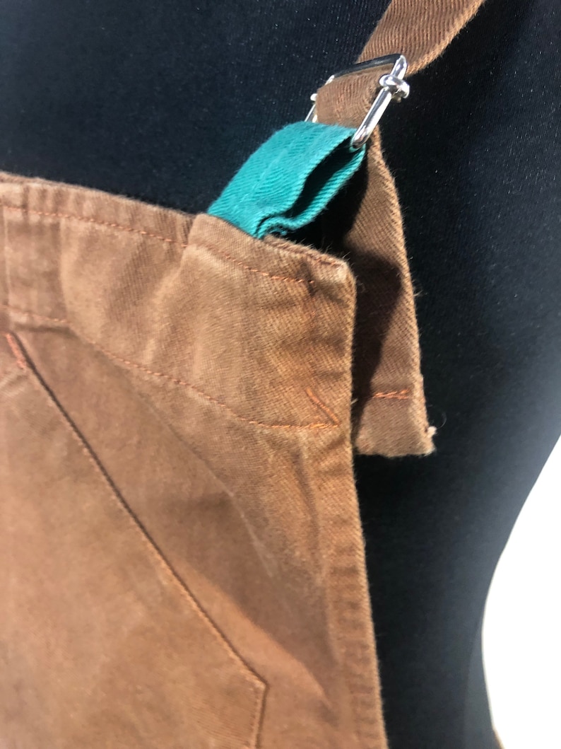 Pode incluir: Close-up de um avental marrom com detalhe de al&ccedil;a verde-azulado e ferragens de metal prateado. O avental tem um bolso e &eacute; feito de um tecido dur&aacute;vel. As al&ccedil;as s&atilde;o ajust&aacute;veis.
