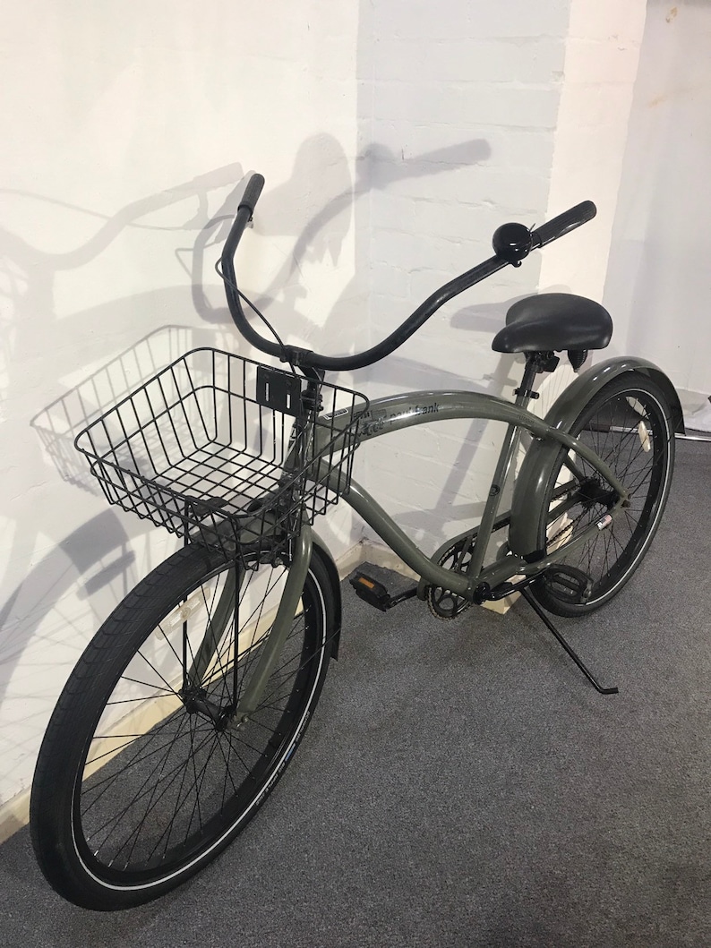 May include: A grey cruiser bicycle with a black wire basket, black tyres, and a black seat. The handlebars curve upwards. The bicycle is leaning against a white wall.