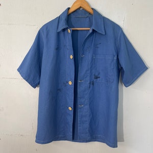Può includere: Una camicia da lavoro blu a maniche corte, con colletto e taschino sul petto. La camicia è chiusa con quattro bottoni rotondi colorati e appesa a una gruccia di legno. Le cuciture visibili dettagliano le cuciture.