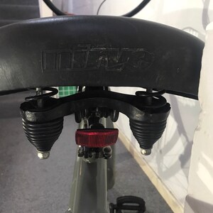 May include: A close-up shot of a black bicycle saddle with the embossed brand name "mitze". The seat is attached to a grey frame, with visible springs and a red reflector. The image is taken from a low angle.