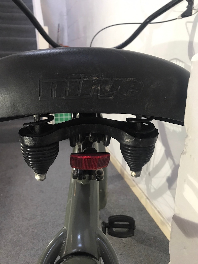 May include: A close-up shot of a black bicycle saddle with the embossed brand name "mitze". The seat is attached to a grey frame, with visible springs and a red reflector. The image is taken from a low angle.