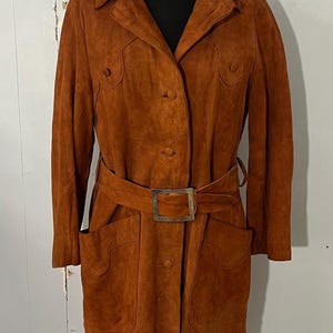 May include: A vintage brown suede coat with a belted waist and large square buckle. The coat features a collared neckline, button closures, and two front pockets. The coat is displayed on a black mannequin.