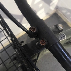 May include: Close-up of a black metal bicycle basket frame with visible rust on the bolts. The frame is curved and attached to a black metal handlebar. The image shows the details of the basket's mounting hardware.