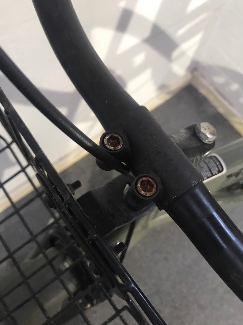 May include: Close-up of a black metal bicycle basket frame with visible rust on the bolts. The frame is curved and attached to a black metal handlebar. The image shows the details of the basket's mounting hardware.