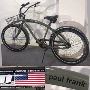May include: An olive green cruiser bicycle with a black basket, seat, and tyres. The bike has the text "paul frank" on the frame and a sticker with the American flag. The bike has a curved handlebar and mudguards.