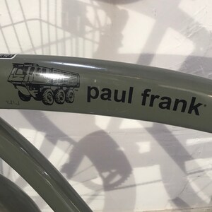 May include: A section of a green bicycle frame with the text "paul frank" in black. A black and white graphic of a dump truck is on the frame. The bicycle is against a white wall.