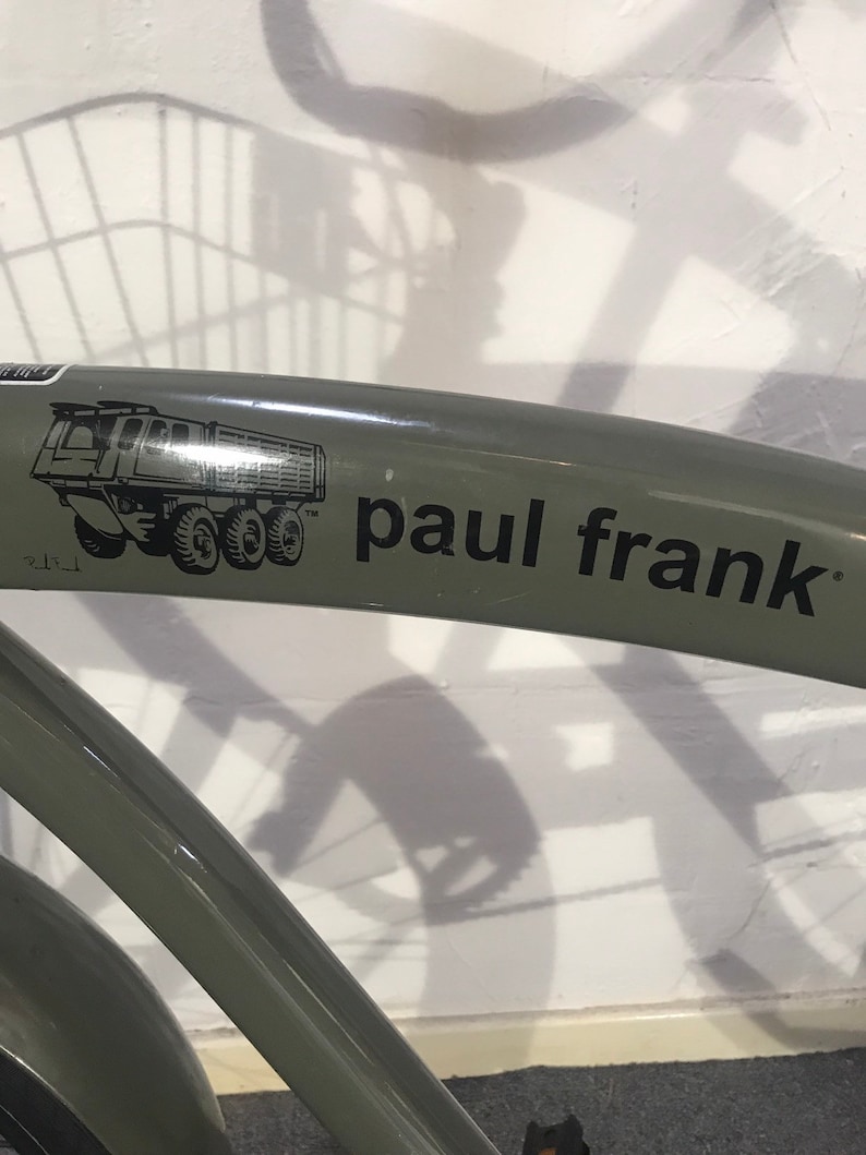May include: A section of a green bicycle frame with the text "paul frank" in black. A black and white graphic of a dump truck is on the frame. The bicycle is against a white wall.