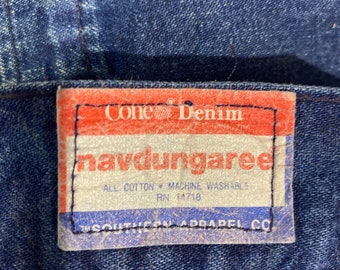 May include: A red and white label sewn onto a blue denim fabric. The label reads "Cone Denim" and "navdungaree". Below the text, it says "All Cotton - Machine Washable" and "RN 14718". The bottom of the label reads "Southern Apparel Co."