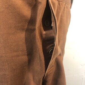 Pode incluir: Close-up de tecido castanho com um bolso e dois bot&otilde;es. O bolso est&aacute; aberto, revelando o interior. O tecido tem uma apar&ecirc;ncia texturizada e os bot&otilde;es s&atilde;o de cor castanha a condizer. A pe&ccedil;a de vestu&aacute;rio parece ser feita de um material dur&aacute;vel.