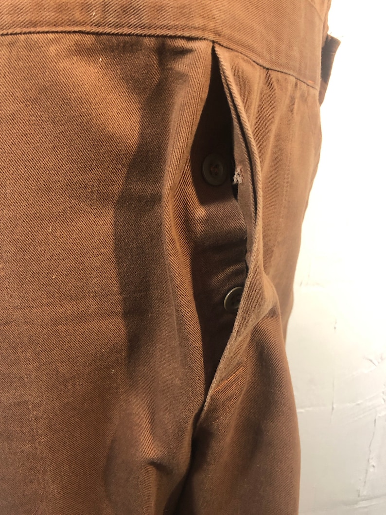 Pode incluir: Close-up de tecido castanho com um bolso e dois bot&otilde;es. O bolso est&aacute; aberto, revelando o interior. O tecido tem uma apar&ecirc;ncia texturizada e os bot&otilde;es s&atilde;o de cor castanha a condizer. A pe&ccedil;a de vestu&aacute;rio parece ser feita de um material dur&aacute;vel.