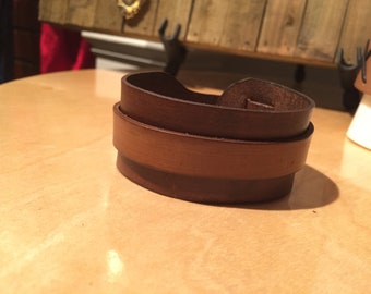 May include: A dark brown leather cuff bracelet featuring a smooth, layered wrap-around design. The leather displays a natural grain texture with varying shades of brown, showing a visible overlapping strap construction secured with a metal stud fastener.