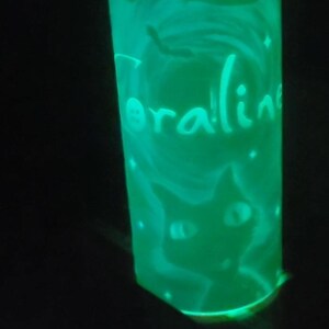 May include: A green glow-in-the-dark glass with a black cat and a character from the movie Coraline. The glass has the word "Coraline" written on it.