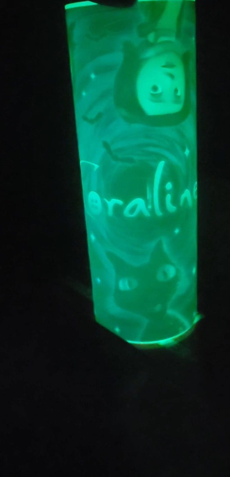May include: A green glow-in-the-dark glass with a black cat and a character from the movie Coraline. The glass has the word "Coraline" written on it.