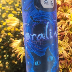 May include: A blue and black tumbler with a graphic of a girl with black hair and a black cat. The girl is wearing a red jacket and the cat has blue eyes. The tumbler has the word "Coraline" written on it with a button.