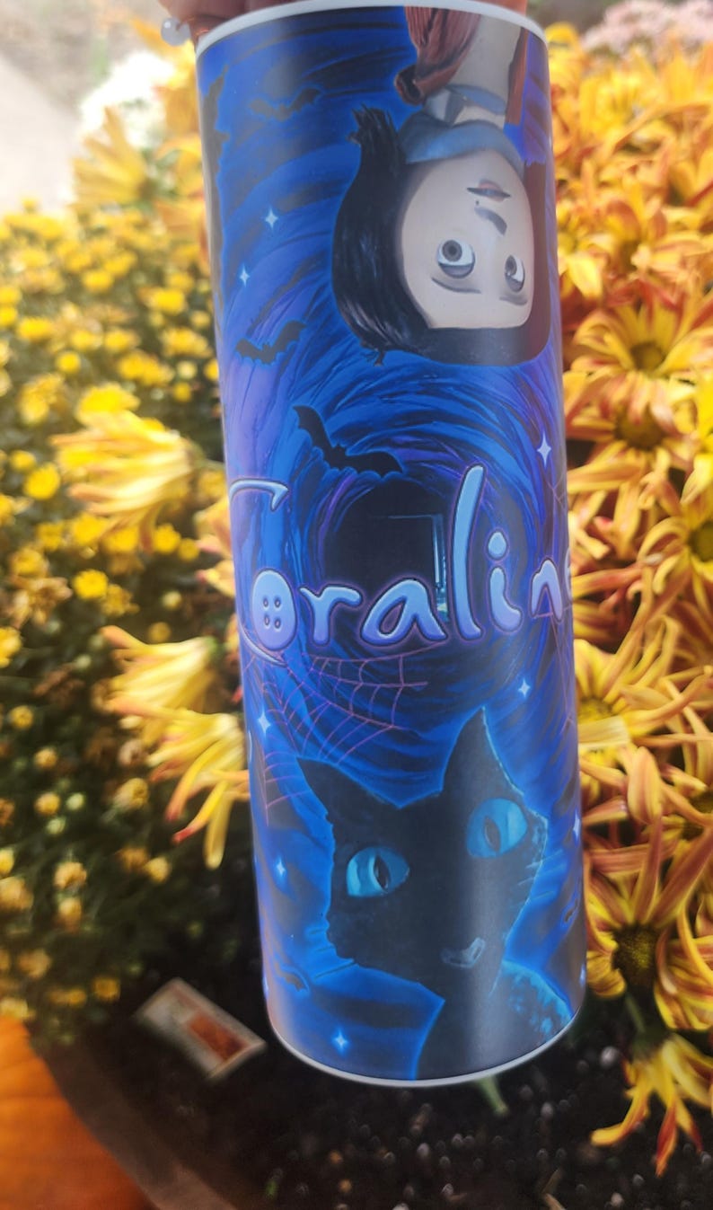 May include: A blue and black tumbler with a graphic of a girl with black hair and a black cat. The girl is wearing a red jacket and the cat has blue eyes. The tumbler has the word "Coraline" written on it with a button.