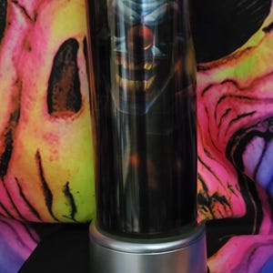 May include: A stainless steel tumbler with a black and white image of a clown with a sinister smile. The tumbler has a silver base.