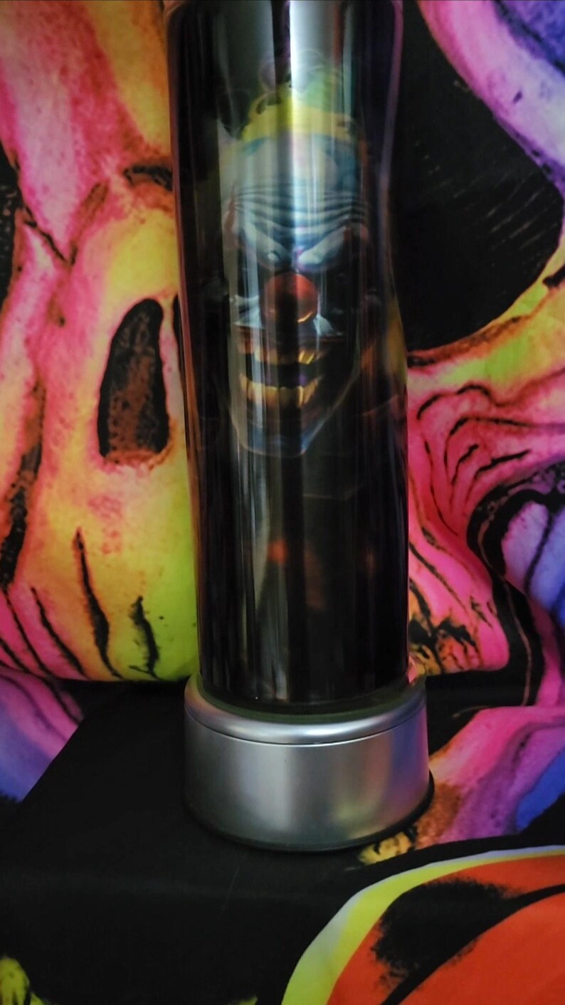 May include: A stainless steel tumbler with a black and white image of a clown with a sinister smile. The tumbler has a silver base.