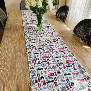 Op de afbeelding: Een wit tafelkleed met een Londens thema, met rode dubbeldekkers, zwarte taxi's en de Union Jack-vlag. De loper ligt op een houten tafel, met een vaas bloemen in het midden. De tafel is ongeveer 200 cm lang.
