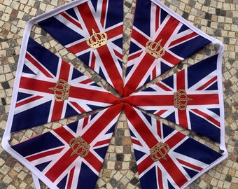 May include: A string of triangular bunting flags featuring the Union Jack design in red, white, and blue. Each flag is adorned with a centered, gold-colored glittery crown emblem. The flags are connected by a white fabric tape along the top edge.