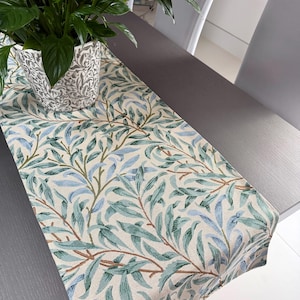 May include: A table runner featuring a botanical design with blue, green, and brown leaves on a cream backdrop. A potted plant with green leaves sits on the runner. The runner is on a dark grey table.