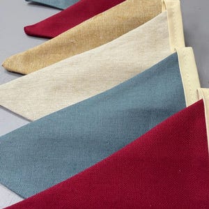 May include: A close-up of a row of colourful triangular fabric pennants. The pennants are in shades of red, blue, beige, and cream. Each pennant has a cream-coloured border. The fabric has a textured appearance.