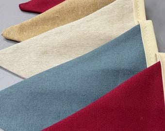 May include: A close-up of a row of colourful triangular fabric pennants. The pennants are in shades of red, blue, beige, and cream. Each pennant has a cream-coloured border. The fabric has a textured appearance.
