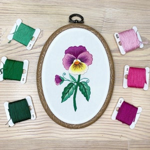 May include: Oval embroidery hoop with a pansy design in purple, yellow, and green. The embroidery is set on white fabric, within a wooden frame. Several spools of green and pink embroidery thread are placed around the hoop, adding a craft theme.