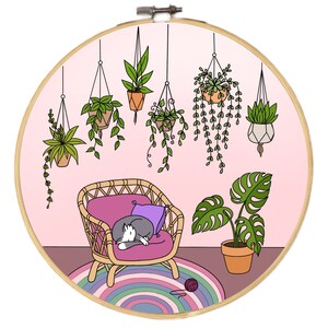 Op de afbeelding: Een ronde borduurring met een gezellige interieurscène. Een kat slaapt op een roze stoel, omringd door hangplanten en een Monstera in pot. Een kleurrijk tapijt en een bol wol maken de illustratie compleet, tegen een roze achtergrond.