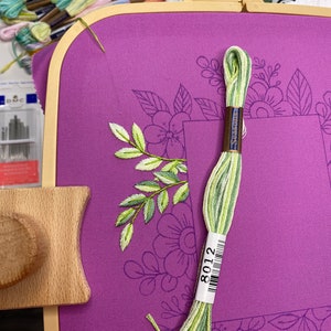 Op de afbeelding: Een paarse borduurring met een bloemenontwerp, met een gedeeltelijk geborduurd blad- en bloemmotief. Een streng groen en geel borduurgaren, met het label 8012, ligt op de stof. Andere garenklosjes en naalden zijn op de achtergrond.