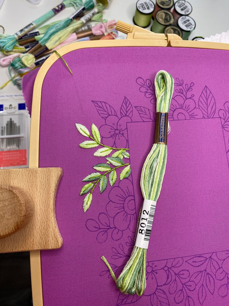 Op de afbeelding: Een paarse borduurring met een bloemenontwerp, met een gedeeltelijk geborduurd blad- en bloemmotief. Een streng groen en geel borduurgaren, met het label 8012, ligt op de stof. Andere garenklosjes en naalden zijn op de achtergrond.