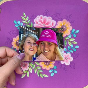 Op de afbeelding: Een paarse borduurring met een bloemenontwerp in roze, geel en teal. Een foto van twee lachende vrouwen wordt voor de borduursel gehouden. Een vrouw draagt een roze hoed met het woord "KALUJH".