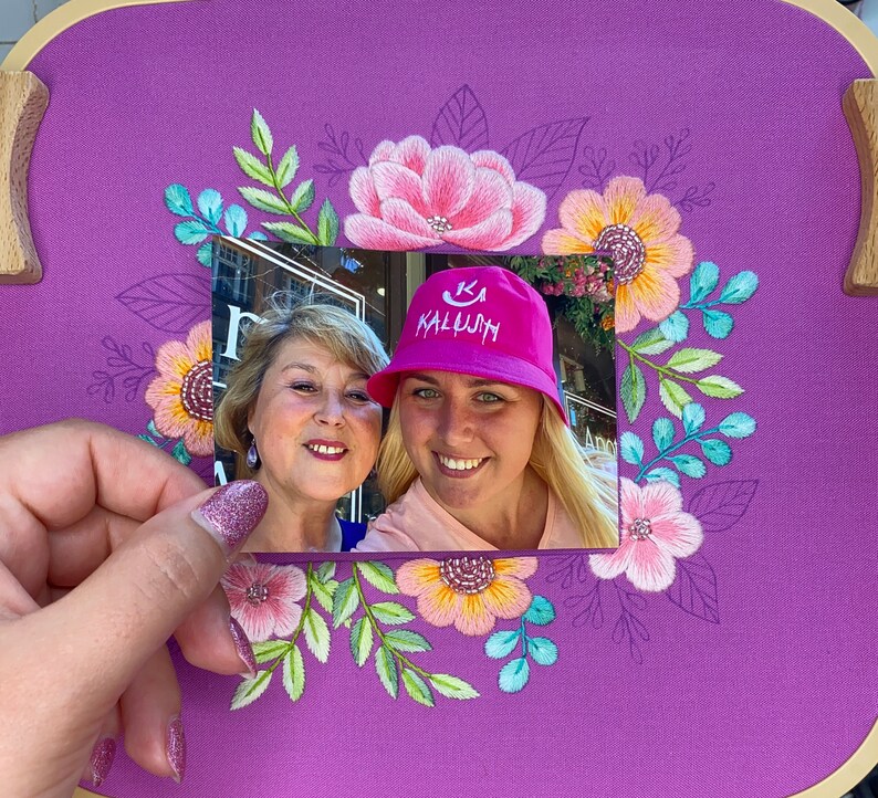 Op de afbeelding: Een paarse borduurring met een bloemenontwerp in roze, geel en teal. Een foto van twee lachende vrouwen wordt voor de borduursel gehouden. Een vrouw draagt een roze hoed met het woord "KALUJH".