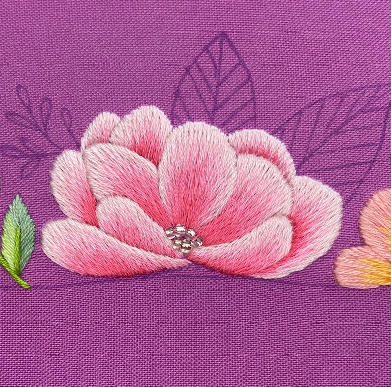 Op de afbeelding: Close-up van een handgeborduurde roze bloem met een cluster van zilveren kralen in het midden, tegen een paarse stoffen achtergrond. De bloemblaadjes zijn beschaduwd met lichtroze en witte draad. Groene en paarse bladercontouren zijn ook zichtbaar.