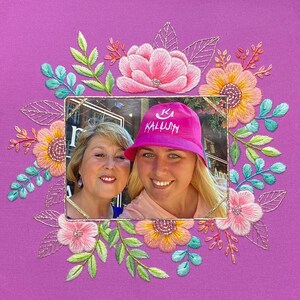 Op de afbeelding: Een ingelijste foto van twee lachende vrouwen, omringd door een geborduurde bloemenkrans. De krans heeft roze, oranje en gele bloemen met groene en blauwe bladeren, alles op een paarse achtergrond. E&eacute;n vrouw draagt een roze hoed met het woord "KALUJH".