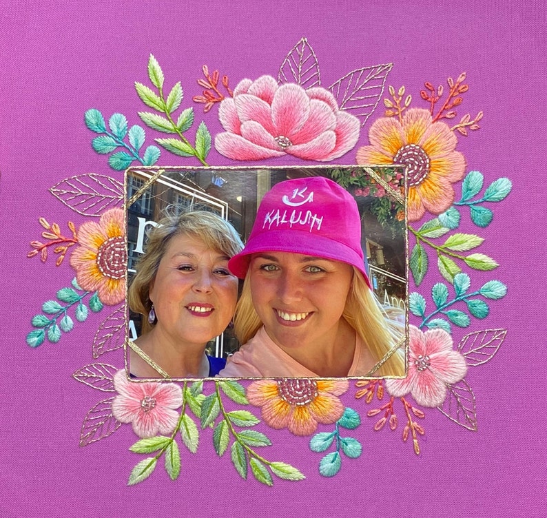 Op de afbeelding: Een ingelijste foto van twee lachende vrouwen, omringd door een geborduurde bloemenkrans. De krans heeft roze, oranje en gele bloemen met groene en blauwe bladeren, alles op een paarse achtergrond. E&eacute;n vrouw draagt een roze hoed met het woord "KALUJH".