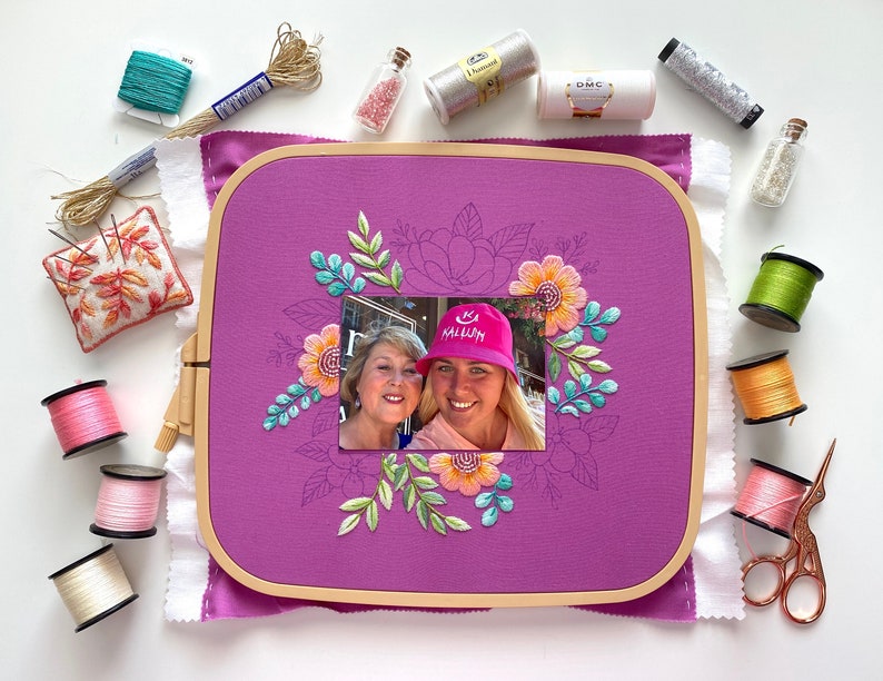 Op de afbeelding: Een paarse borduurring met een foto van twee vrouwen, omringd door bloemenborduurwerk in oranje, turquoise en groen. Het beeld wordt omringd door spoelen garen, naalden en scharen, wat duidt op een knutselproject.