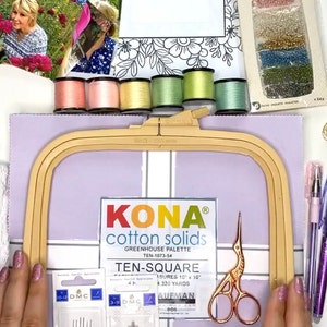Op de afbeelding: Een platte lay-out van knutselbenodigdheden met borduurgaren, naalden, een schaar, een borduurring en stof. De afbeelding bevat een verpakking glaskralen, een bloemenontwerp en een kleurenkaart. De items zijn gerangschikt op een lichtpaars oppervlak.