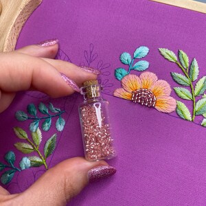 Op de afbeelding: Een kleine glazen fles gevuld met roze kralen, gehouden voor een paarse stof met geborduurde bloemen en bladeren. Het borduurwerk heeft tinten roze, oranje, groen en blauw. De fles heeft een kurk.